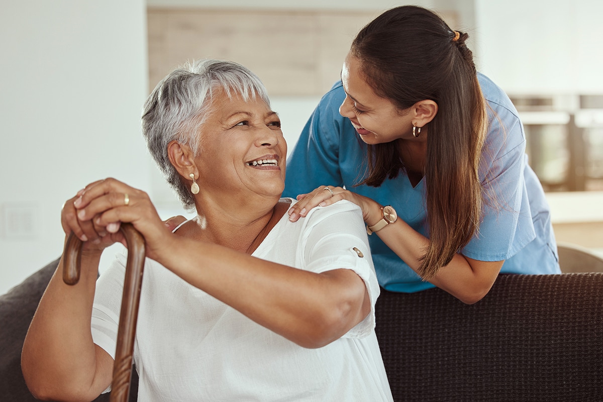 Happy, relax and senior woman with caregiver smile while sitting on a living room sofa in a nursing home. Support, help and professional nurse or healthcare worker helping elderly lady or patient.