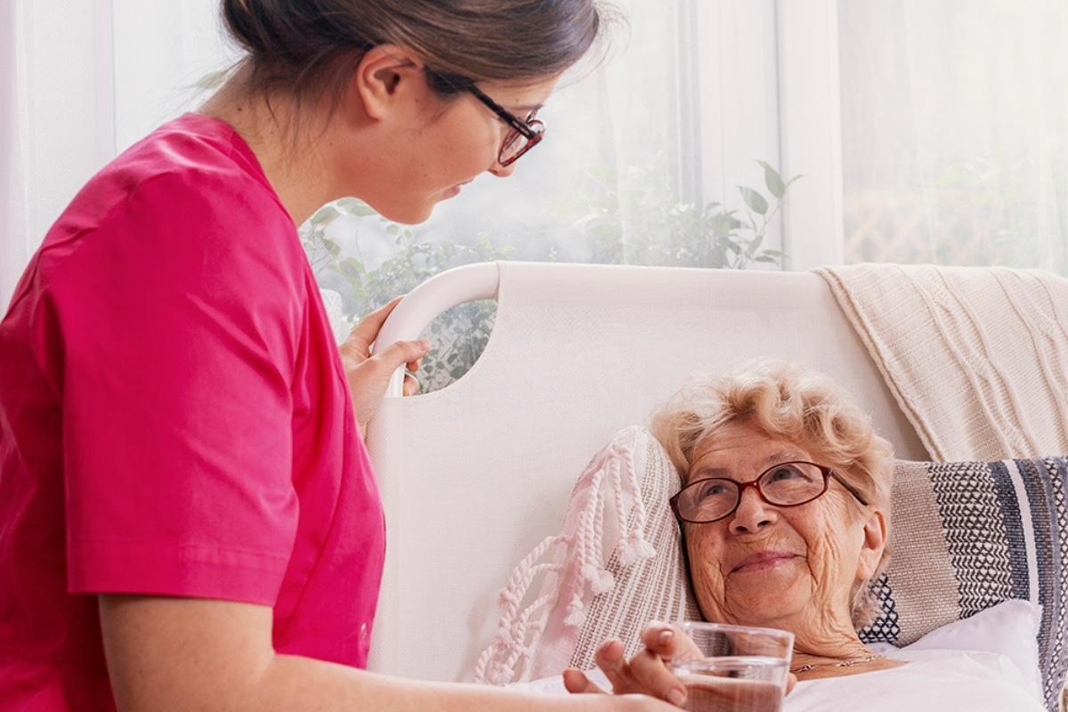 Caregiver offering a glass of water to an older woman resting in bed